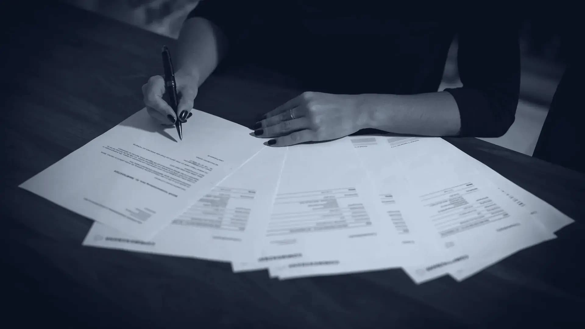 Hands working through a spread of printed forms on a dark desk